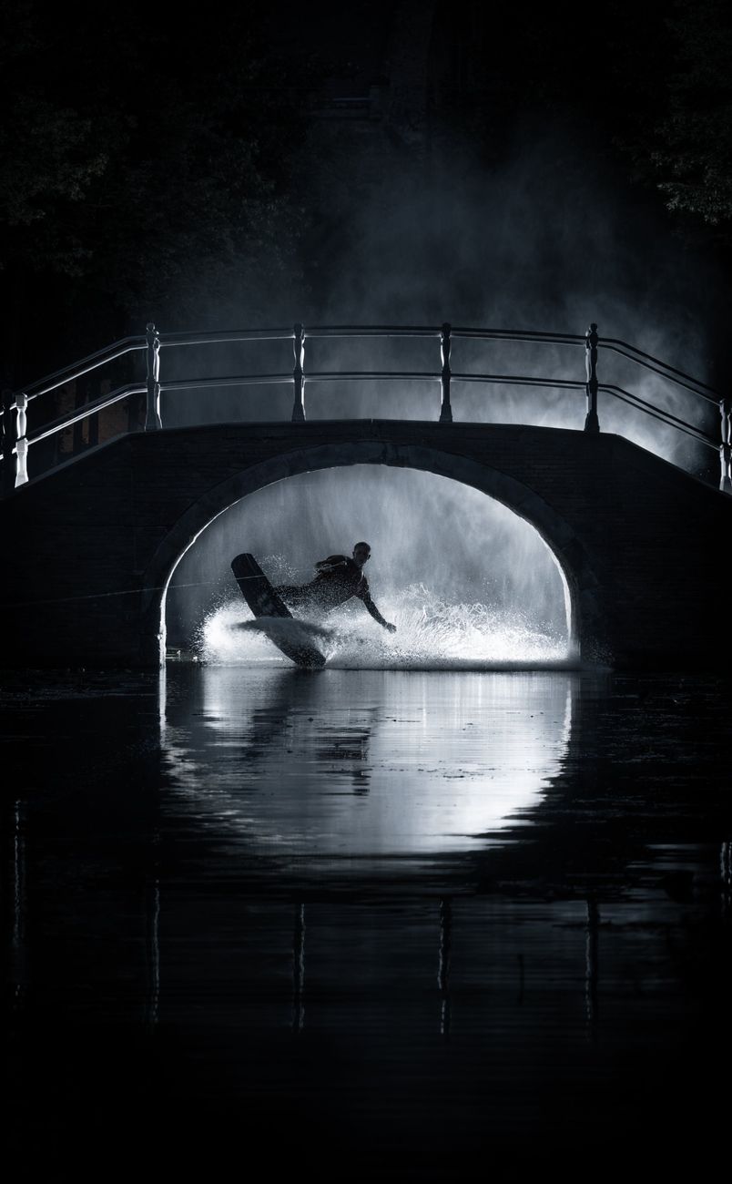 Wakeboarder spraying water under a lit canal bridge at night